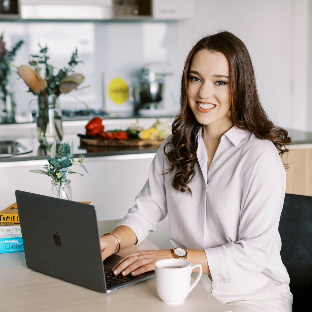A smiling woman sitting at a kitchen counter working on a laptop with a cup of coffee and a vibrant kitchen setting in the background.
