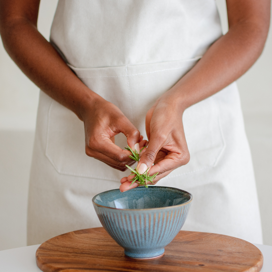 A person in a white apron preparing food by placing herbs in a blue bowl on a wooden table.