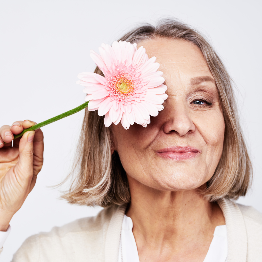 Older woman smiling and holding a pink flower over one eye.
